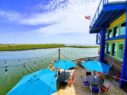 A vibrant waterfront view featuring blue umbrellas, a patio, and a building with large windows beside a calm waterway.