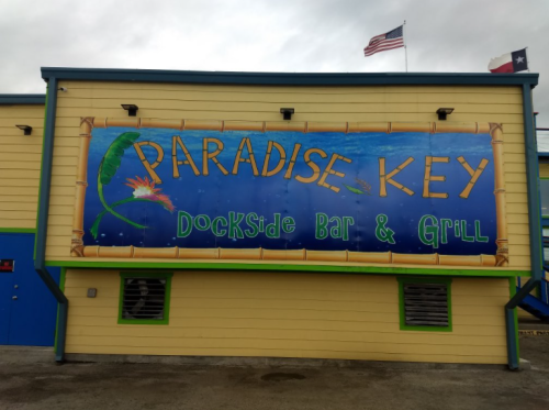 Colorful sign for "Paradise Key Dockside Bar & Grill" on a yellow building, with flags in the background.