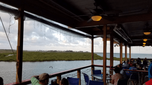 View from a waterfront restaurant with diners, overlooking a calm waterway and marshland under a cloudy sky.