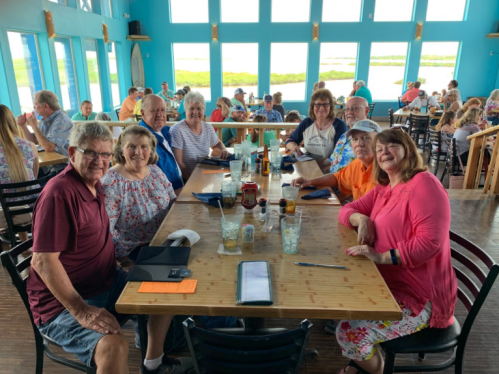A group of eight people smiling around a table in a bright restaurant with large windows and a scenic view.