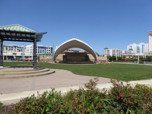 A large outdoor stage with a curved roof in a park, surrounded by green grass and modern buildings in the background.