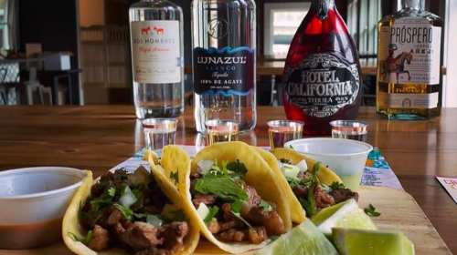 Three tacos filled with meat and toppings, accompanied by tequila bottles and shot glasses on a wooden table.