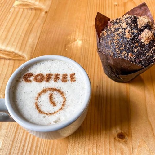 A cup of coffee with "COFFEE" written in cocoa powder beside a chocolate muffin on a wooden table.