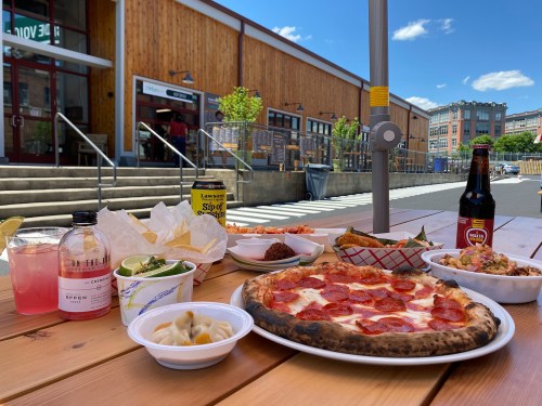 A table outdoors with pizza, drinks, and various dishes, set against a sunny urban backdrop.
