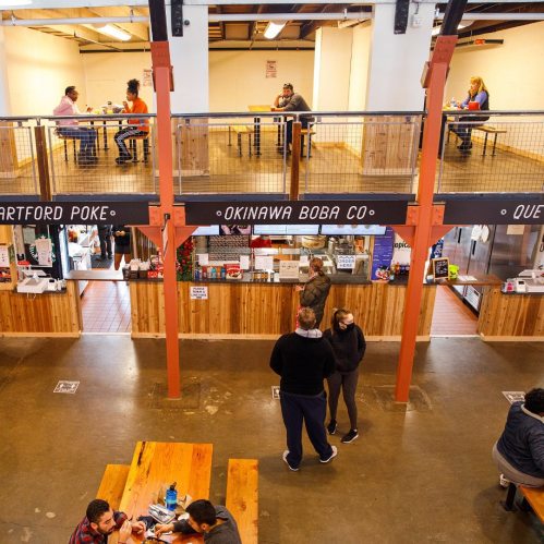 A bustling food hall with people dining at tables and a counter serving various cuisines. Bright and inviting atmosphere.