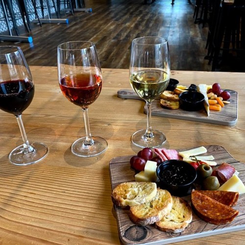 Three glasses of wine beside two wooden boards with cheese, meats, olives, and crackers on a rustic table.