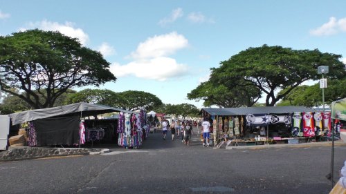 A vibrant outdoor market with stalls selling clothing and crafts, surrounded by lush trees and a clear blue sky.