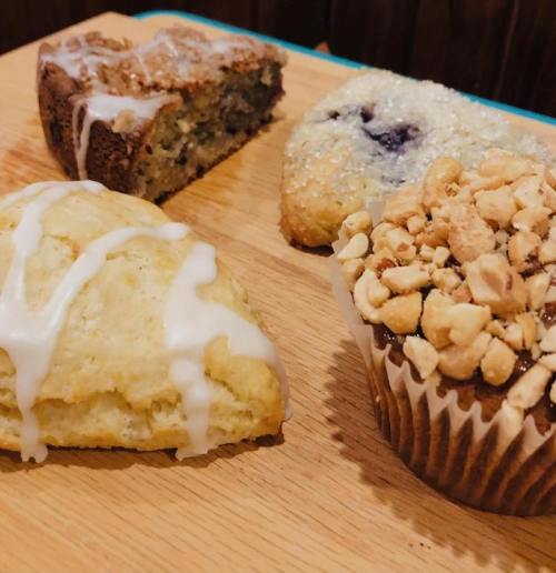 A wooden board displays four baked goods: a scone, a muffin topped with nuts, and two types of sweet bars.