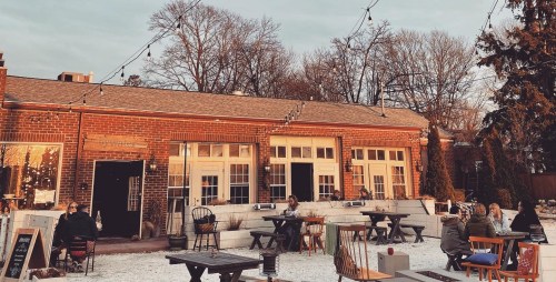 A cozy outdoor dining area with people seated at tables, surrounded by trees and string lights, during sunset.