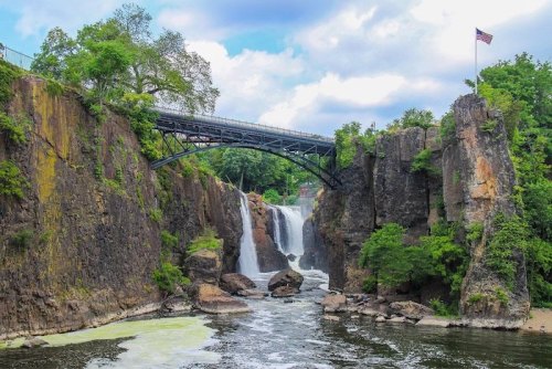 A scenic view of a waterfall cascading between rocky cliffs, with a bridge overhead and a flag in the background.