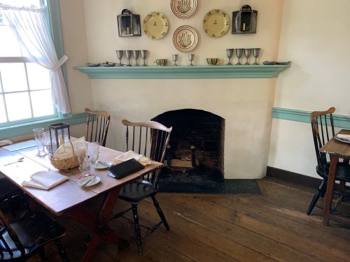 Cozy dining room with wooden tables, chairs, a fireplace, and decorative plates on the wall. Natural light from the window.