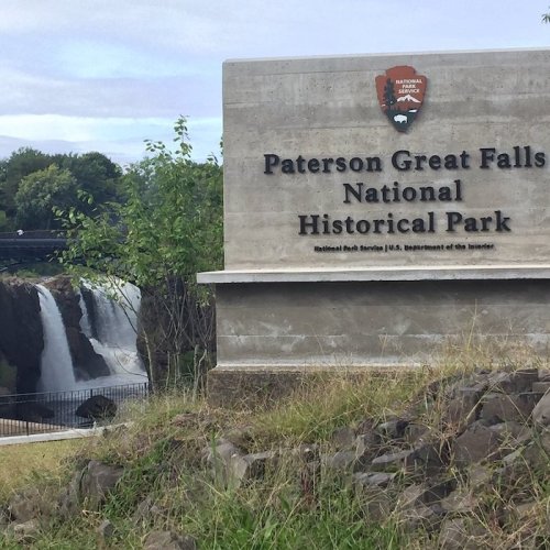 Sign for Paterson Great Falls National Historical Park with waterfalls in the background. Green grass and trees visible.