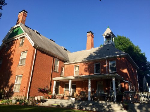 A large red brick house with a porch, multiple chimneys, and a tower, set against a clear blue sky.