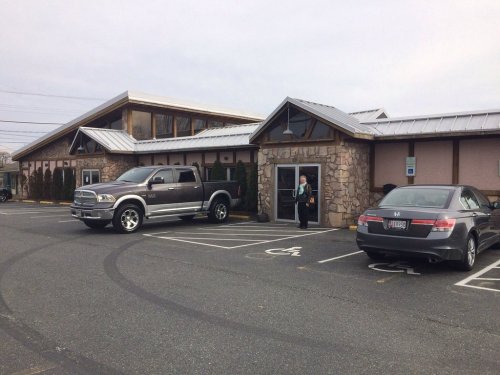 A gray pickup truck and a black sedan parked outside a stone building with a sloped roof on a cloudy day.