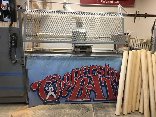 A woodworking machine for crafting baseball bats, with a "Cooperstown Bats" banner and unfinished wooden bat blanks nearby.