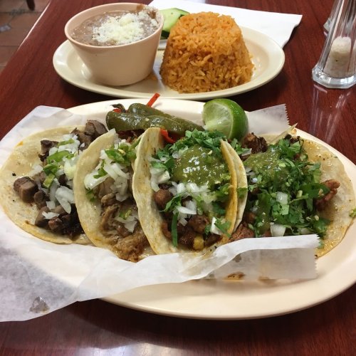 A plate of three tacos topped with onions and cilantro, served with rice, beans, and a lime wedge.