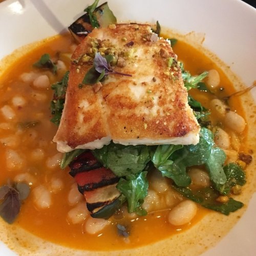 A plate featuring grilled fish on a bed of greens and white beans in a vibrant orange sauce, garnished with herbs.