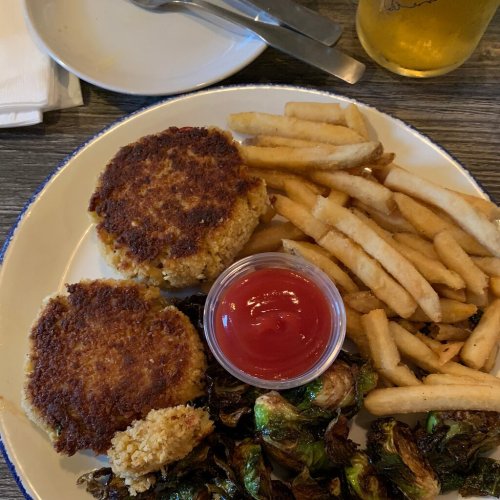 A plate with two golden crab cakes, fries, roasted Brussels sprouts, and a small cup of ketchup.