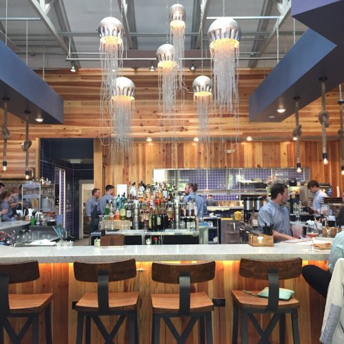A modern bar with wooden accents, hanging jellyfish lights, and a well-stocked liquor shelf in a lively restaurant setting.