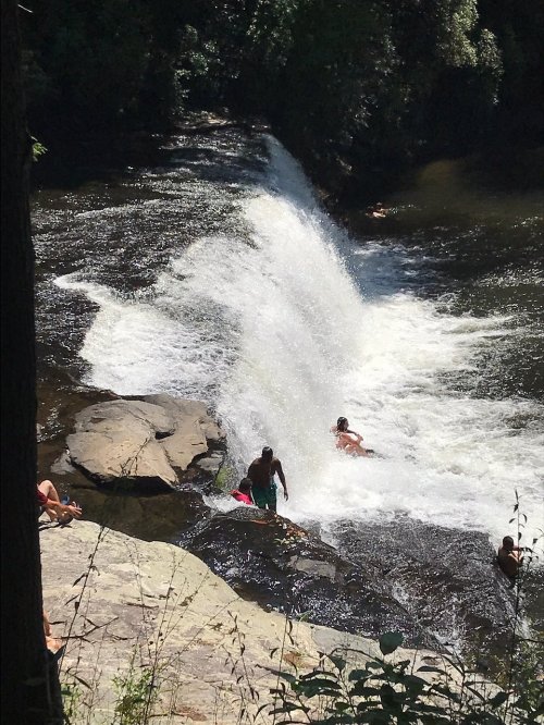 A waterfall cascades into a river, with people enjoying the water and relaxing on the rocks nearby.