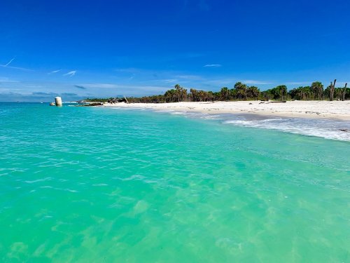 A serene beach with turquoise water, white sand, and a clear blue sky, lined with palm trees in the background.