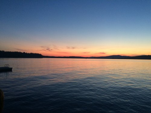 A serene sunset over a calm lake, with silhouettes of distant hills and a clear blue sky transitioning to warm hues.