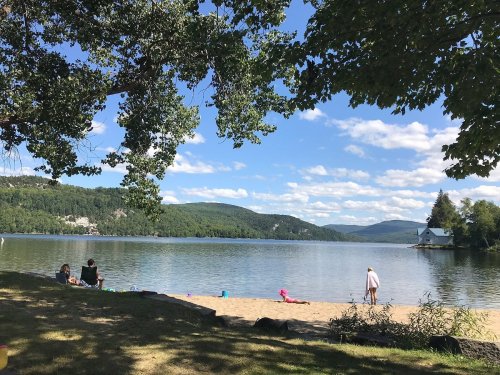 A serene lake scene with people on the sandy shore, surrounded by trees and mountains under a blue sky with fluffy clouds.