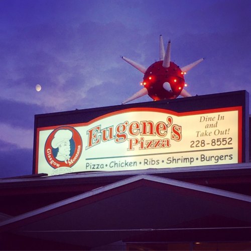 Sign for Eugene's Pizza featuring a chef graphic, with the moon visible in a twilight sky.