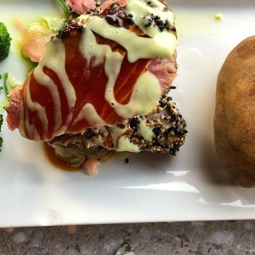 A plate featuring sesame-crusted fish drizzled with sauce, accompanied by a baked potato and broccoli.