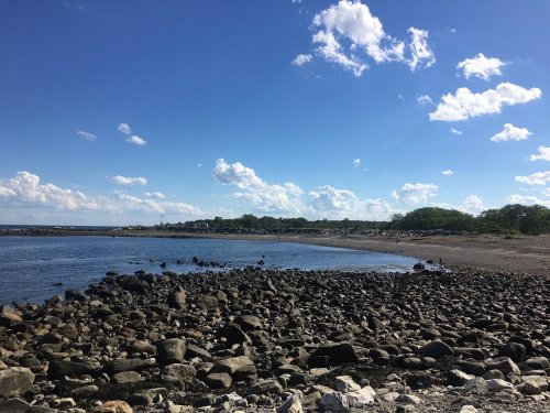 A rocky beach under a blue sky with scattered clouds, featuring calm water and greenery in the background.