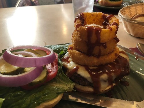 A stacked burger with cheese, barbecue sauce, and onion rings, served with lettuce, tomato, and pickles on a plate.