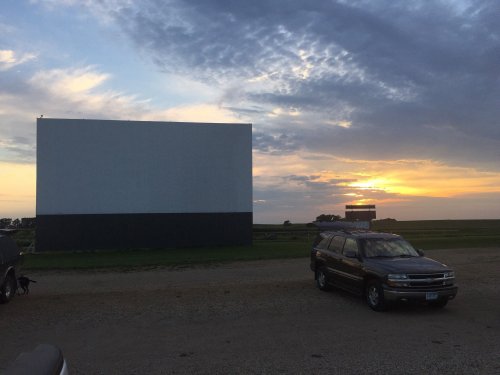 A sunset view at a drive-in theater, featuring a blank movie screen and parked cars in the foreground.