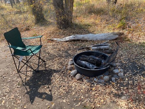A camping scene featuring a green chair next to a fire pit surrounded by stones and logs in a wooded area.