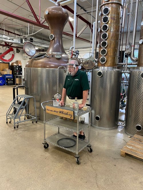 A man stands by a cart in a distillery, with large copper and stainless steel stills in the background.