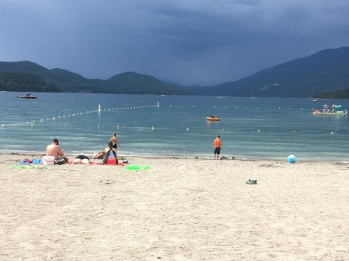 A sandy beach with people relaxing, boats on the water, and dark clouds looming over distant mountains.