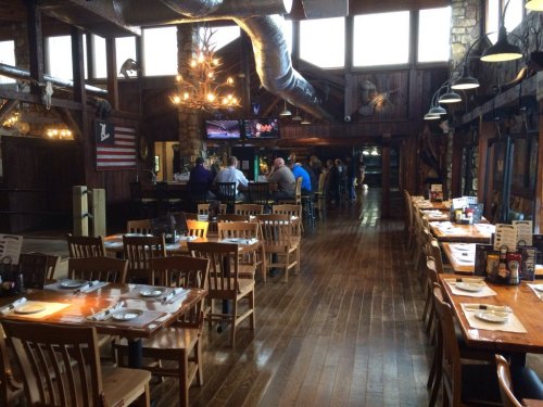 Interior of a rustic restaurant with wooden tables, chairs, and a bar area, featuring large windows and a cozy atmosphere.