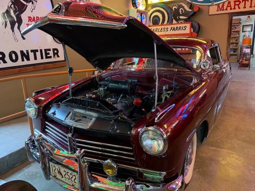 A vintage maroon car with its hood open, showcasing the engine, in a retro-themed setting with neon signs.