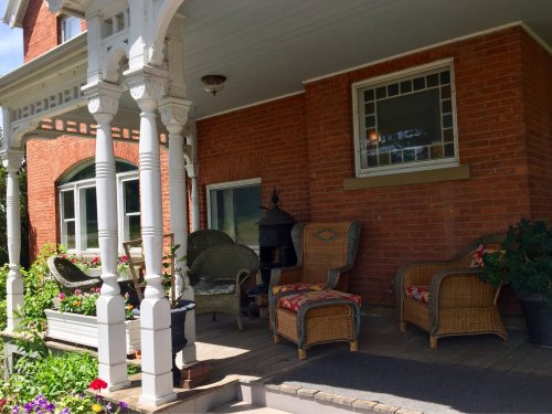 A cozy porch with wicker chairs, colorful cushions, and potted plants, framed by white columns and red brick walls.