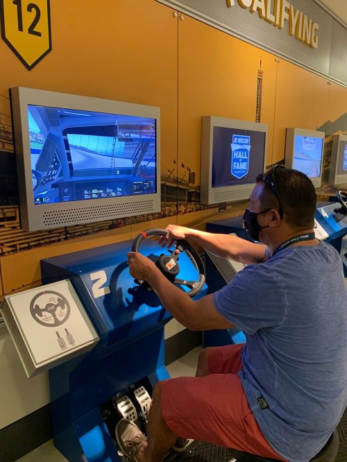 A person wearing a mask is sitting at a racing simulator, gripping the steering wheel and focused on the screen.