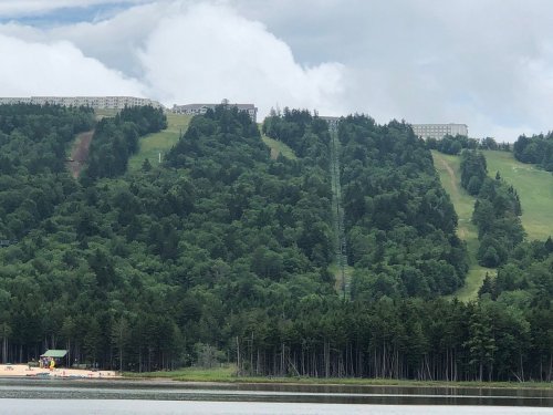 A lush green hillside with a ski slope, a building at the top, and a sandy area near a lake in the foreground.