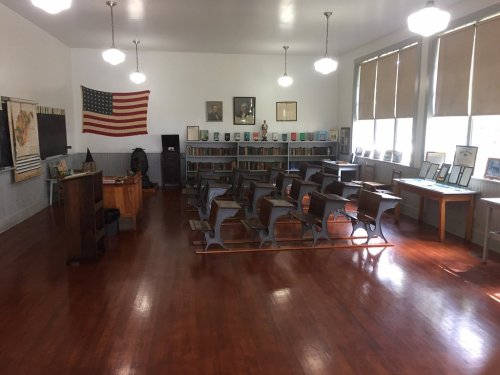 A vintage classroom with wooden desks, bookshelves, an American flag, and historical portraits on the walls.