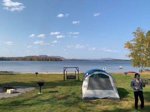 A serene lakeside scene with a tent, picnic area, and a person walking, surrounded by trees and mountains in the background.