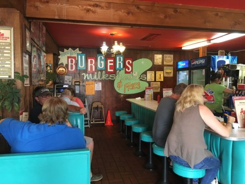 A cozy diner interior with teal seating, a "Burgers, Milkshakes & Fries" sign, and patrons enjoying their meals.