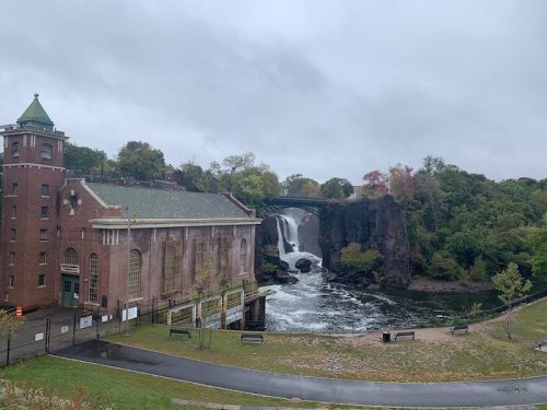 Historic building beside a waterfall and river, with a bridge in the background, under a cloudy sky.