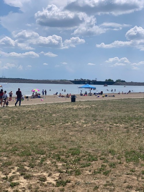 A sandy beach scene with people sunbathing, swimming, and enjoying the water under a partly cloudy sky.