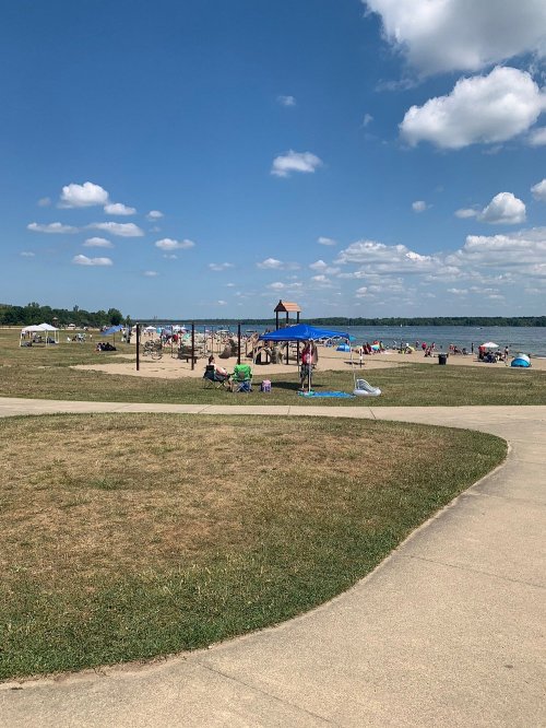 A sunny beach scene with people relaxing on the sand, playground equipment, and blue skies with fluffy clouds.