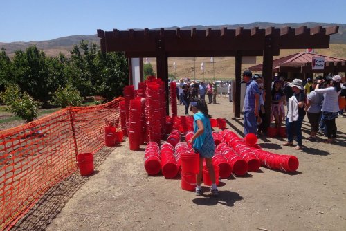 A child picks up a red bucket near a stack of buckets at a busy outdoor event with people in the background.