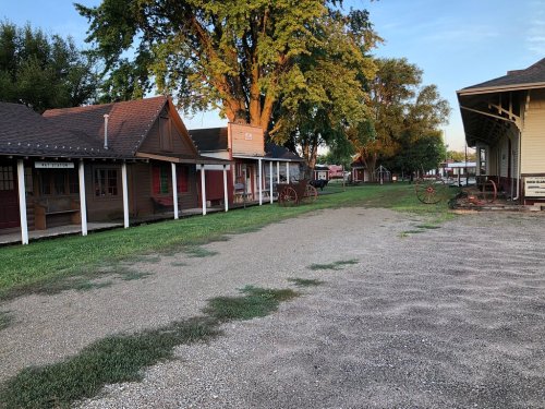 A quiet street lined with rustic buildings and wagons, surrounded by greenery and trees in the early morning light.
