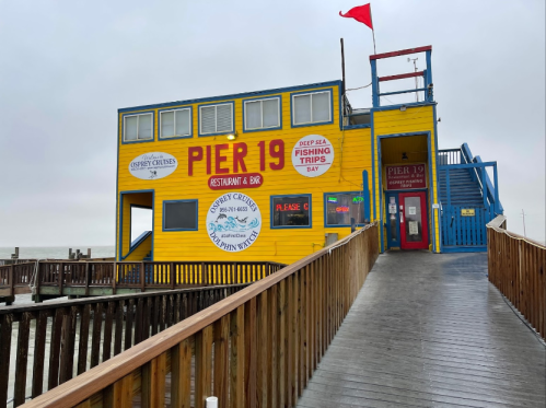 Colorful two-story building labeled "Pier 19" with a red flag, wooden walkway, and signs for fishing trips and dining.