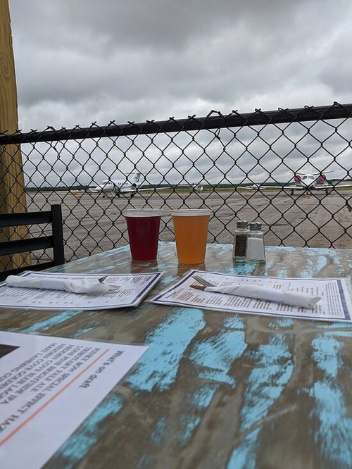 Two drinks on a table with menus, overlooking an airport runway through a chain-link fence on a cloudy day.